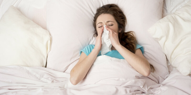 women with allergies in blue shirt laying in white bed blowing nose
