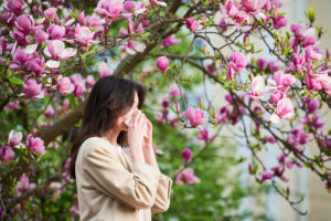 Young woman sneezing and blowing nose with nasal handkerchief in front of blooming tree looking for the Best Allergists in CT