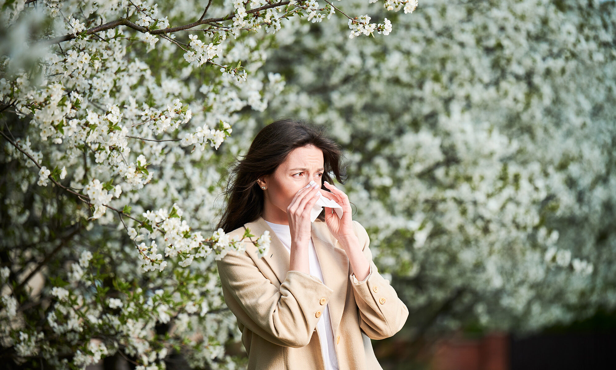 A woman blows her nose under a tree while searching for the best allergy doctors in danbury ct